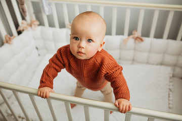 Beautiful little girl in a crib in a cozy light room in a sweater of fashionable shades. learns to stand at the support. cheerful little girl standing in the crib at home