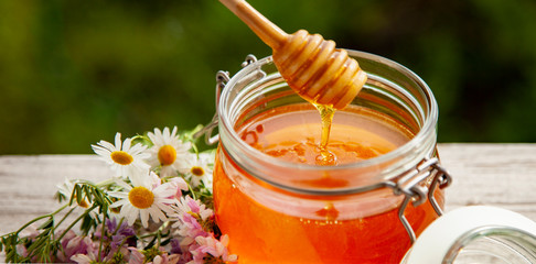 Honey in glass jar with bee flying and flowers on a wooden floor.