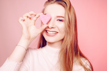 Obraz na płótnie Canvas Valentine's Day, a girl on a pink background, closes her eye with a heart. The blonde smiles beautifully, laughs, love. March 8, gifts for the girl.