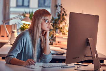 Blond serious woman in glasses is working on computer while sitting in the office.