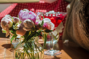 Beautiful little bouquet of undiscovered white pink peonies against the background of a red drink in a can. Buds of flowers stand in a glass little jar on the table in the sunlight, casting a shadow.