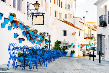 Fototapeta na wymiar Mijas Pueblo Blanco, charming small village, picturesque empty street in old town with bright blue tables chairs of local cafe, flower pots hanging on white washed houses walls, Costa del Sol, Spain