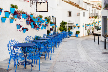 Mijas Pueblo Blanco, charming small village, picturesque empty street in old town with bright blue tables chairs of local cafe, flower pots hanging on white washed houses walls, Costa del Sol, Spain