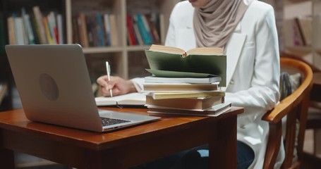 Muslim student is doing research at desk full of books in library. Girl wearing hijab is preparing for exam or making a project, then looking at camera 4k - Powered by Adobe