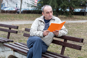 Fototapeta premium Middle Age Man Sitting on Bench in Public Park and Reading Book