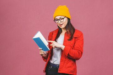 Beautiful young woman wearing casual and eyeglasses standing isolated over pink background, reading a book.