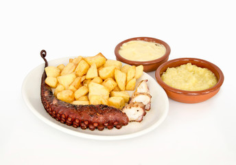 Plato con pulpo a la brasa con patatas y dos cazuelas de barro con distintas salsas sobre fondo blanco