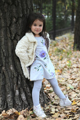 A little girl with long dark hair in a fluffy white coat and colorful dress is standing in front of a big tree. Blurred park background