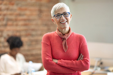 Pretty older business woman, successful confidence with arms crossed in financial building. Cheerful attractive businesswoman crossing arms on chest and looking at camera. - obrazy, fototapety, plakaty
