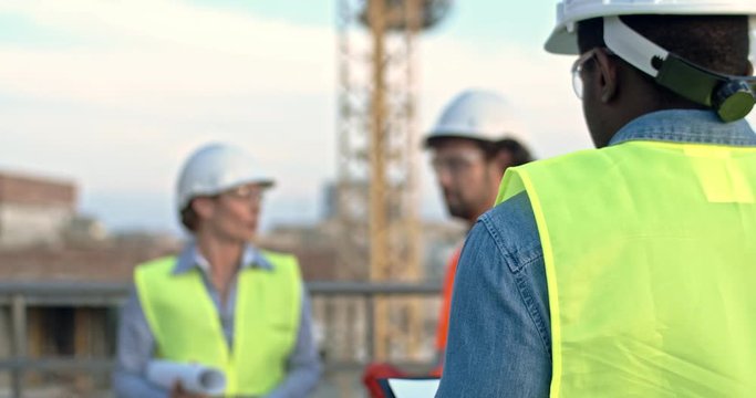 View from the back on the African American man builder in hardhat coming to his Caucasian colleagues - man and woman while they talking outdoor at the constructing site. Rear.