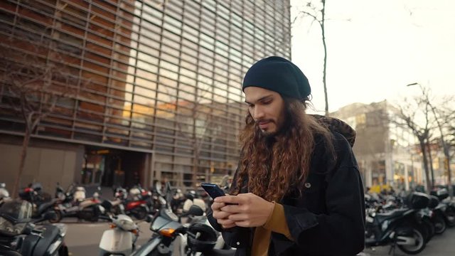 Portrait of long haired hipster walking on street in modern city in slow motion. He wears black jacket with leopard hoodie. He is typing on a cell phone.