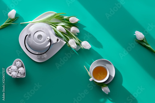 Spring geometric flat lay. Tea cup, pot, sweets and white tulips on green. Valentine's day, Mother's day, international women day March 8. Diagonal sunlight, long shadows, modern design, top view