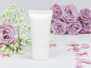White plastic cosmetic tube on a white background with flowers roses and petals, mockup, beauty product and spa concept