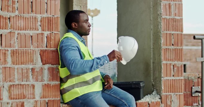 African American young man builder sitting at the building site and resting while taking off hardhat and relaxing. Outside.