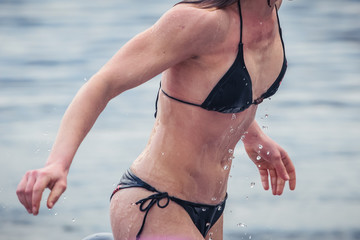 Unknown woman in black bikini getting out of the cold water in winter time, with drops falling from...