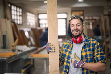 Portrait of handsome smiling carpenter with wood material at workshop holding thumbs up. Do it...