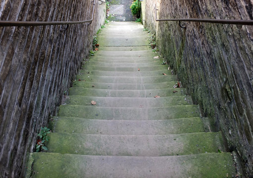 a perspective view of narrow old outdoor stone steps descending between walls with weeds and moss growing in the corners