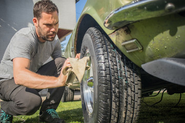 Young man with a hip beard cleaning a wheel of an old green vintage car on a home lawn. Cleaning...