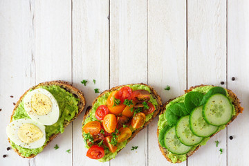 Healthy avocado toast assortment with eggs, tomatoes and cucumber spinach. Bottom border flat lay over a white wood background.
