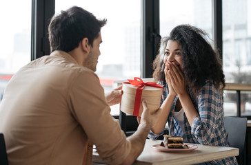 Romantic man giving birthday gift to beautiful emotional woman. Portrait of lovely multiracial couple sitting together in cafe, dating. Valentines day concept