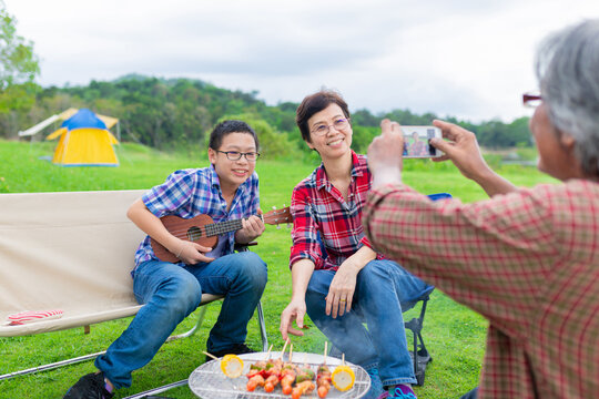 close up asian female, old asian man use smartphone take a photo to family, they travel and picnic in lakeside, they camping and grilling barbecues with stove, happiness family activity 