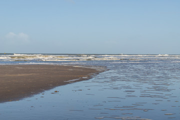 Dutch coastline. The sea surf at the sand and waves coming in with sea foam. Blue sky on a winter day.