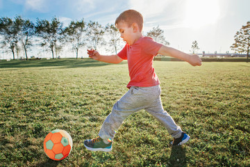 Fototapeta premium Little preschool Caucasian boy playing soccer football on playground outside. Kid kicking hitting ball. Happy authentic candid childhood lifestyle. Seasonal summer outdoors activity for children.