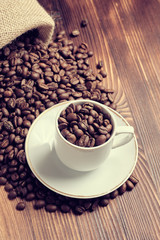 A white cup on coffee beans from a knitted bag with a burnt wooden background. Vertical. Toned.