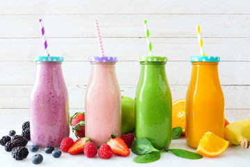 Various healthy smoothies in bottles with ingredients. Side view against a white wood background. Blueberry, strawberry, green and orange.