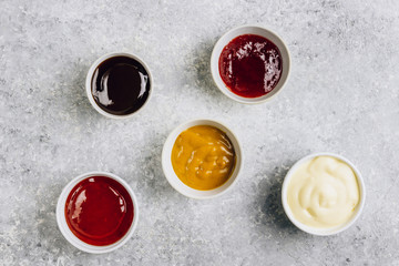 Obraz na płótnie Canvas White bowls of various dip sauces on gray background. Mustard, ketchup, cheese, teriyaki and cranberries sauces. Top view. Flat lay