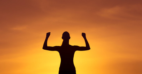 Strong healthy active female silhouette flexing on sunset sky 