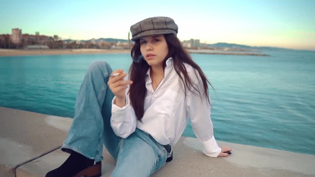 Portrait of young beautiful stylish woman in a white shirt and grey cap smoking on sea terrace with ocean view.