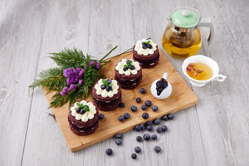Row of four brown cakes with blue berry on top placing together violate flower, tea pot and berry as decorated item on the wooden plate