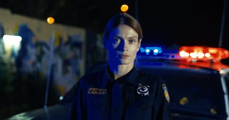 Portrait of attractive Caucasian young female police officer looking in car at night. Policewoman at the police car withh lights outdoor in dark.