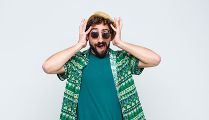 young tourist man raising hands to head, open-mouthed, feeling extremely lucky, surprised, excited and happy against white wall