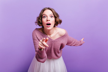Cute girl with pink makeup expressing amazement and pointing finger. Indoor photo of curly shocked...