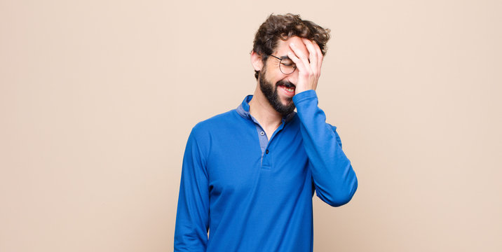 young handsome man laughing and slapping forehead like saying d’oh! I forgot or that was a stupid mistake against flat wall