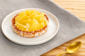 Small pineapple tart or tartlet with glazed pineapple fruit pieces and grated coconut on grey napkin by a golden spoon, over a wooden background.