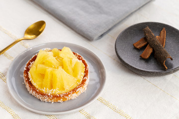Pineapple tart or tartlet with grated coconut by a golden spoon and a small plate with cinnamon sticks on a white and gold table cloth.