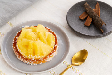 Pineapple tart or tartlet with coconut by a golden spoon and a small plate with cinnamon sticks on a white and gold table cloth.