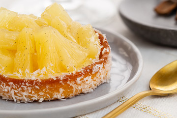 Small pineapple tart or tartlet with glazed pineapple fruit pieces and grated coconut by a golden spoon, over a white and gold table cloth.