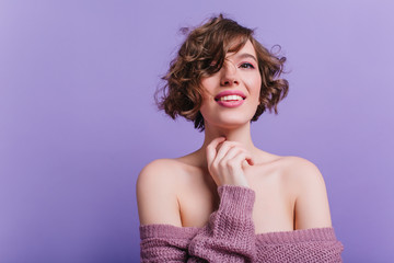 Winsome young woman with cute curly hairstyle smiling on purple background. Studio shot of relaxed brunette girl in woolen sweater.