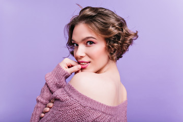 Close-up portrait of interested young woman with short brunette hair wears sweater. Indoor photo of sensual lady playfully looking over shoulder on purple background.