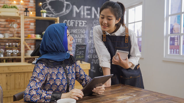 Attractive young waitress take order from muslim woman customer holding digital tablet computer in modern coffee shop. girl barista in apron smiling service islam female client in urban cafe bar