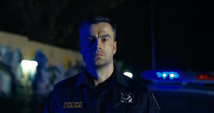 Close up of attractive brave Caucasian police officer turning face to camera and looking straight outdoor. Cop at car with lifgts on street at night. Portrait of policeman.