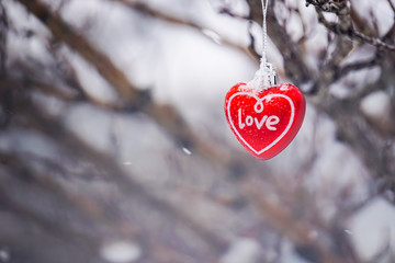 Red heart love ornament for Valentine's Day hanging on outdoor tree during snowfall