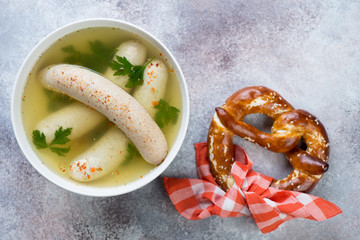 White bowl with boiled munich sausages in a broth and a pretzel, top view on a beige stone background