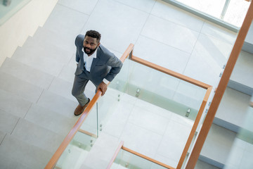 High angle view of serious Afro-American bearded businessman in suit moving up stairs and looking...