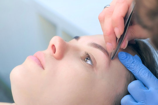Professional beautician plucking eyebrows with tweezers to woman in beauty salon after tint eyebrow procedure, closeup view. Cosmetologist shaping girl's eyebrows in cosmetology clinic.