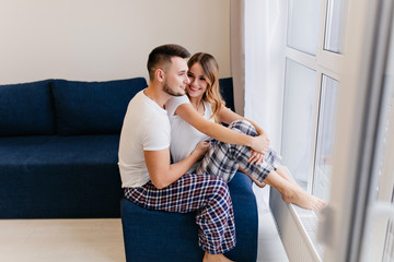 Pleased man in checkered pants looking at window. Charming blonde woman resting at home in good morning.